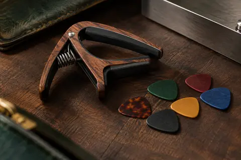 Capodastre à pince au motif bois accompagné de six médiators colorés sur un établi de musicien sombre.