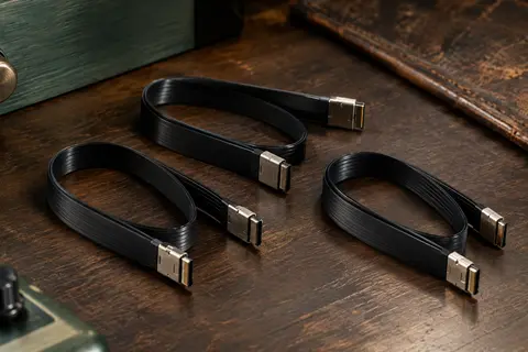 Trois câbles patch plats noirs Ernie Ball avec connecteurs métalliques, disposés sur un établi en bois sombre.