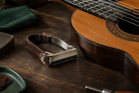 Capodastre plat à lanière nickelé posé sur un établi près d’une guitare classique.