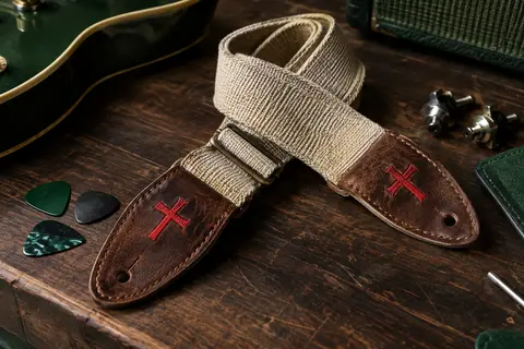 Sangle de guitare en coton avec extrémités en cuir brodées d’une croix rouge, posée sur un établi